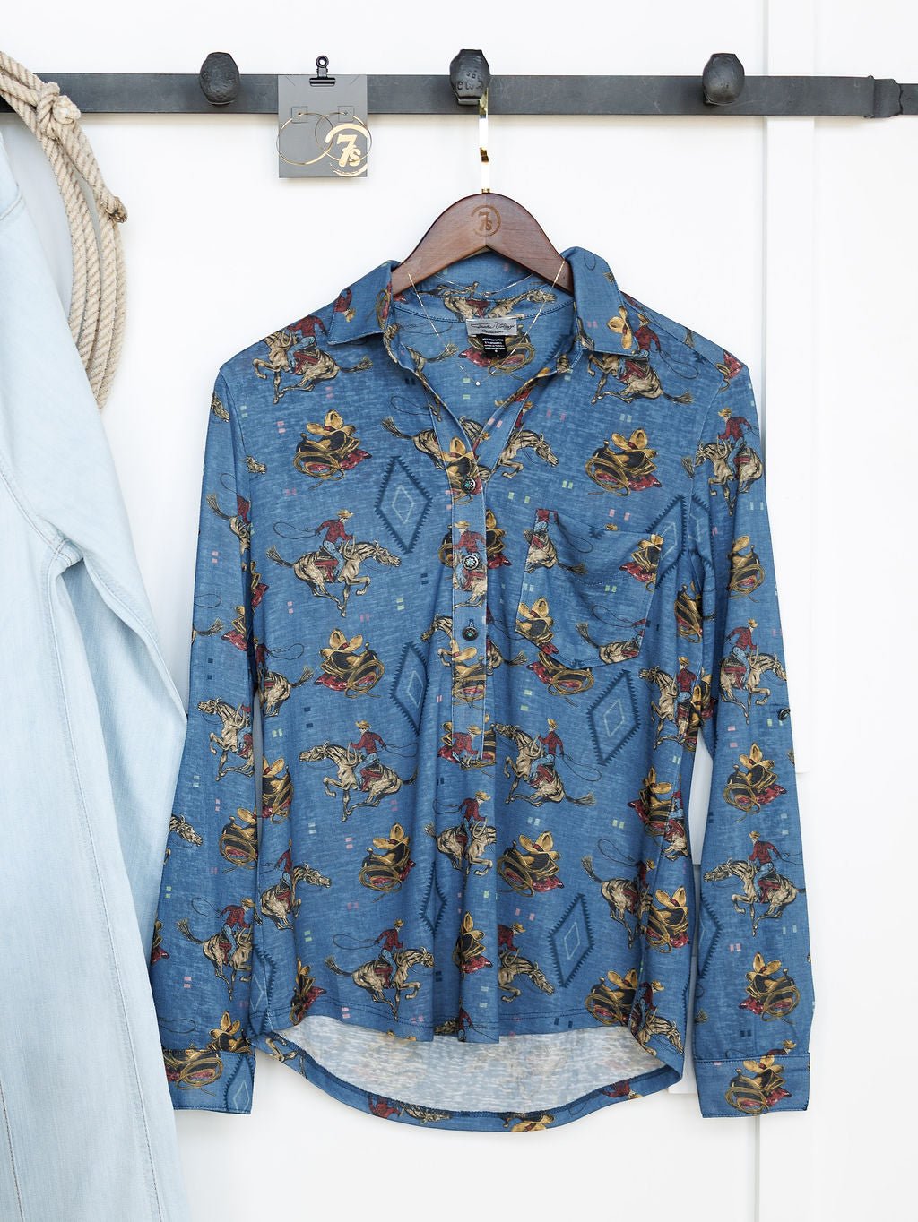 The Paducah Western Print Button Up Top by Savannah Sevens western life{&}style, in steel blue with a cowboy and horse motif, hangs on a wooden hanger beside a light denim jacket. Inspired by Tasha Polizzi.