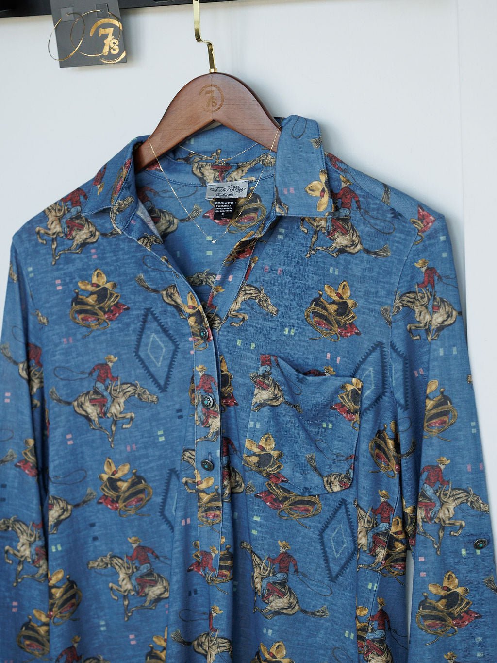The Paducah Western Print Button Up Top by Savannah Sevens western life{&}style features a steel blue color, pocket, and cowboy with horses and hats print. Displayed on a wooden hanger against a white background.