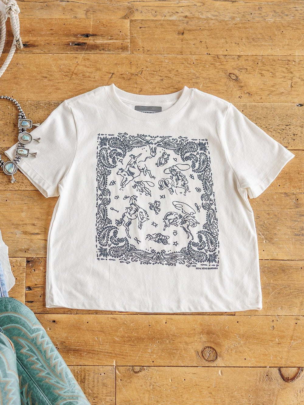 The Paisley Cowboy tee by Savannah Sevens western life{&}style is a white 100% cotton shirt with a black bandana-style print of celestial and animal graphics, shown flat on a wooden floor with jewelry and fabric nearby.