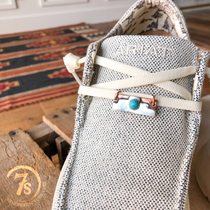 A close-up of a light Ariat shoe with cream laces, featuring a Kingman turquoise and copper pin and a Palco Shoe Tag by Savannah Sevens western life{&}style. A patterned rug, wooden floor, and the 7S logo appear in the background.