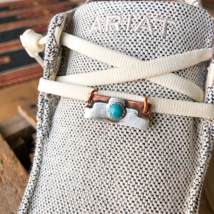 Close-up of a light ARIAT shoe with white laces, featuring the Savannah Sevens western life{&}style Palco Shoe Tag with copper accents and a decorative metal charm set with Kingman turquoise threaded onto the laces.