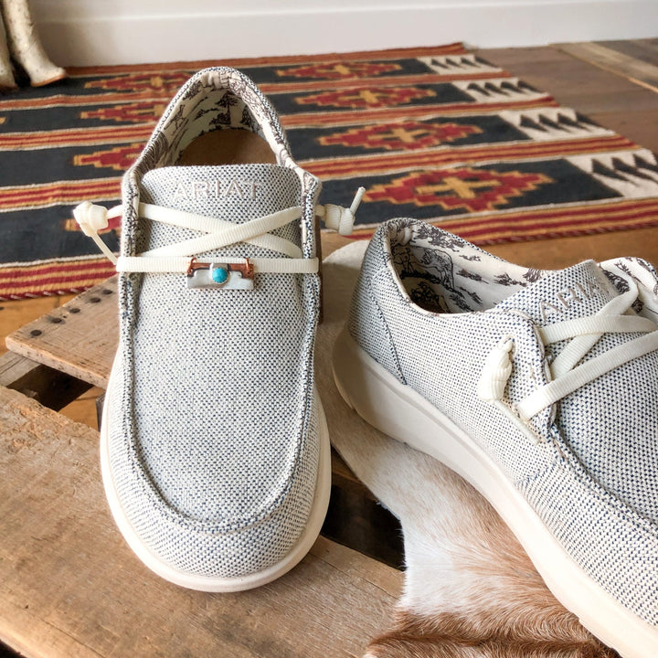 A pair of light gray Palco Shoe Tag shoes by Savannah Sevens western life{&}style with white laces and patterned lining rests on wood, one accented by a sterling silver Kingman turquoise ring; a patterned rug appears behind.