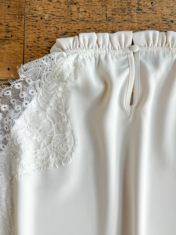 A close-up of the Palisade Cream Lace Trim Top by Savannah Sevens western life{&}style, featuring delicate lace trim on one side, gathered fabric at the top, and a small keyhole opening below the collar, displayed on a wooden surface.