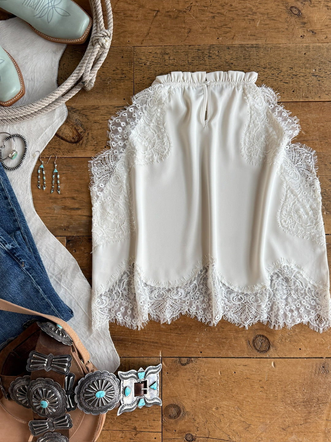 The Palisade Cream Lace Trim Top by Savannah Sevens western life{&}style is displayed on a wooden surface, accompanied by turquoise cowboy boots, turquoise jewelry, a belt with silver and turquoise buckles, and a pair of jeans.