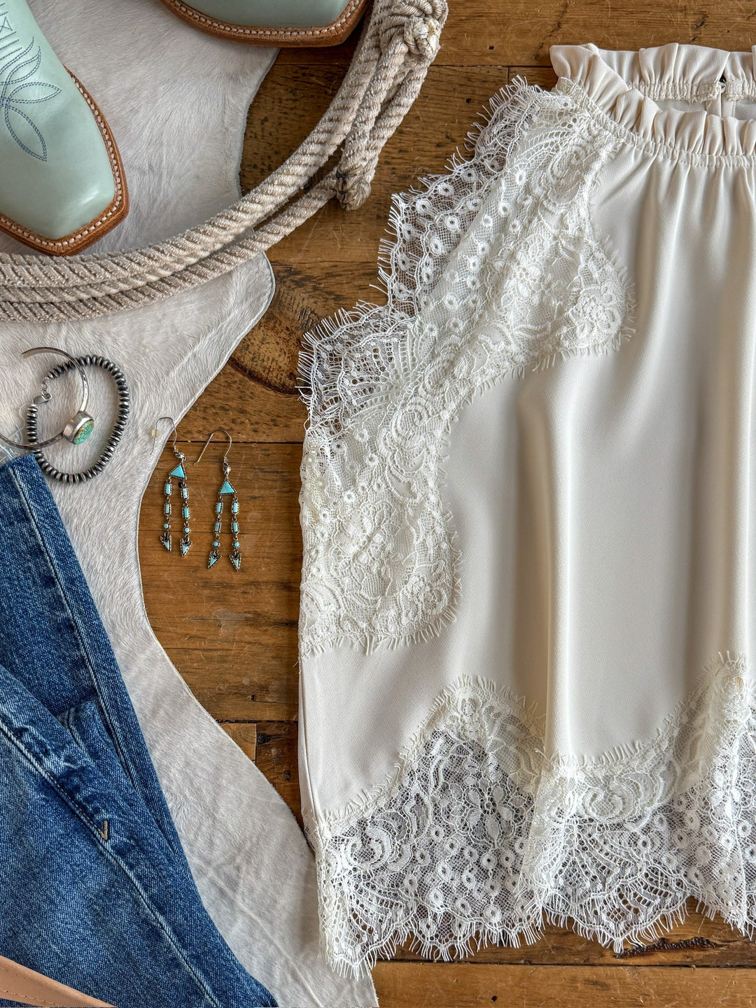 Flat lay of the Savannah Sevens western life{&}style Palisade Cream Lace Trim Top styled with turquoise earrings, denim jeans, silver jewelry, rope, and cowboy boots on a rustic wooden surface.