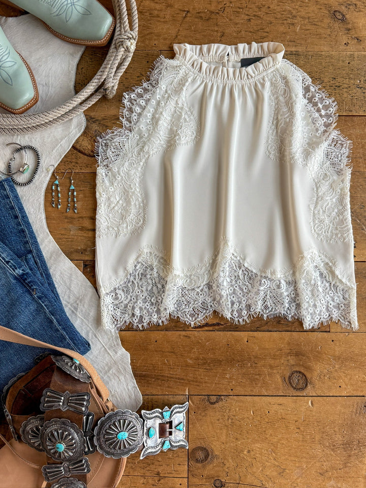The Palisade Cream Lace Trim Top by Savannah Sevens western life{&}style is displayed on a wooden floor, surrounded by turquoise jewelry, a brown belt, blue jeans, a cowboy hat, and turquoise cowboy boots.