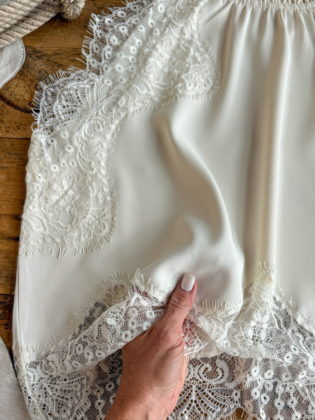 A hand  holds the hem of an ivory silk and lace garment, revealing the intricate details of the Savannah Sevens western life{&}style Palisade Cream Lace Trim Top displayed on a wooden surface.