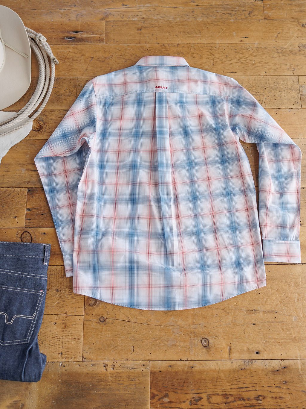 The Palmer Ariat Men's Plaid Button Up by Savannah Sevens western life{&}style, featuring light blue, red, and white plaid with sun protection and moisture-wicking fabric, is laid flat on a wooden floor beside a cowboy hat and folded blue jeans.