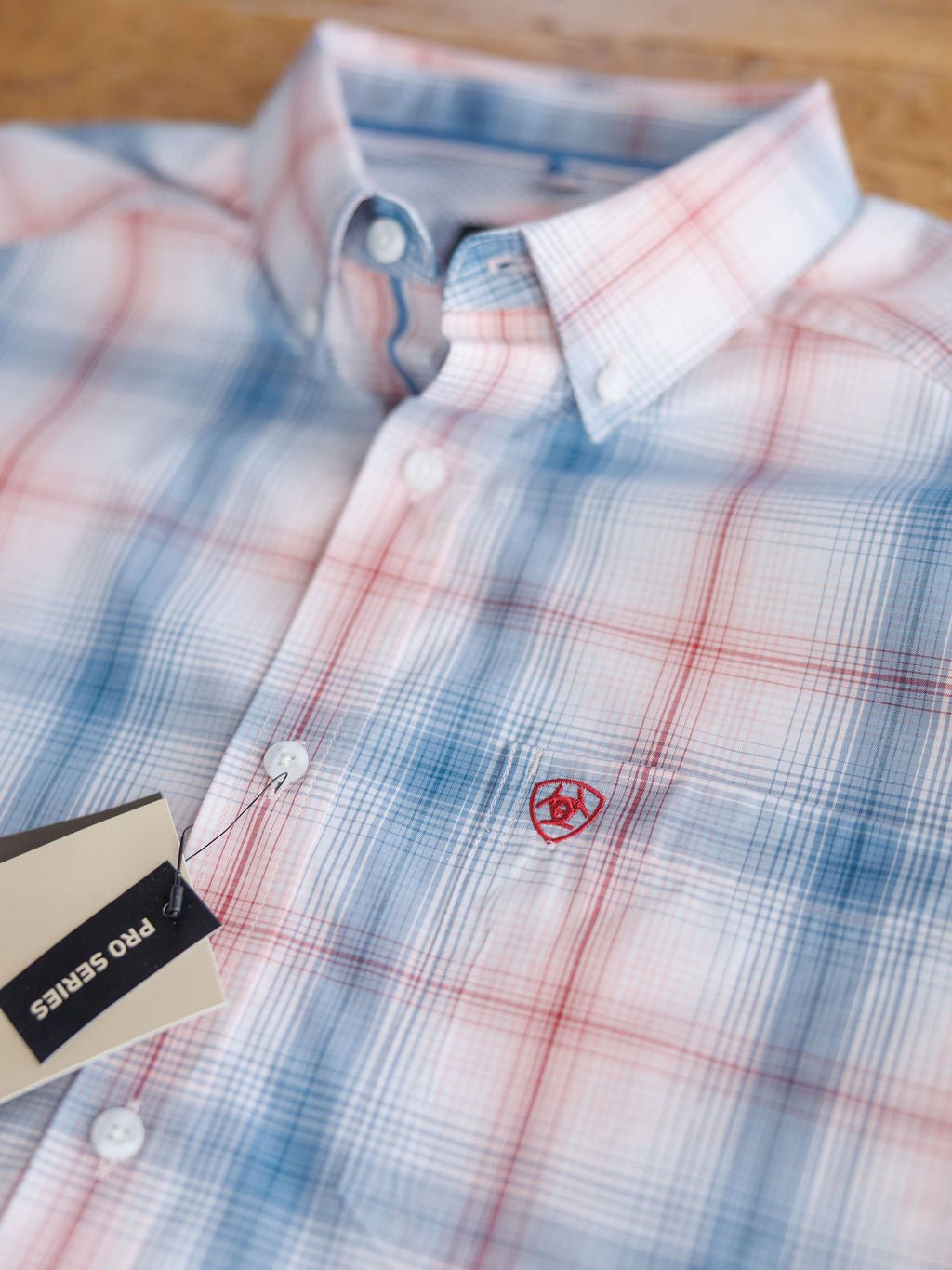 A close-up of the Palmer Ariat Men's Plaid Button Up from Savannah Sevens western life{&}style shows its red and blue plaid, embroidered red chest logo, sun protection features, and a black PRO SERIES tag attached to a button.