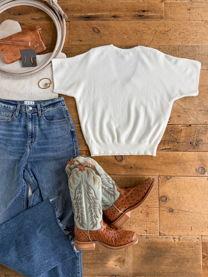 Flat lay of a western outfit featuring the Savannah Sevens western life{&}style Panaca White Knit Top, blue jeans, light brown tooled cowboy boots with green accents, tan belt, leather wallet, gold hoop earrings, and a ring arranged on a wooden floor.