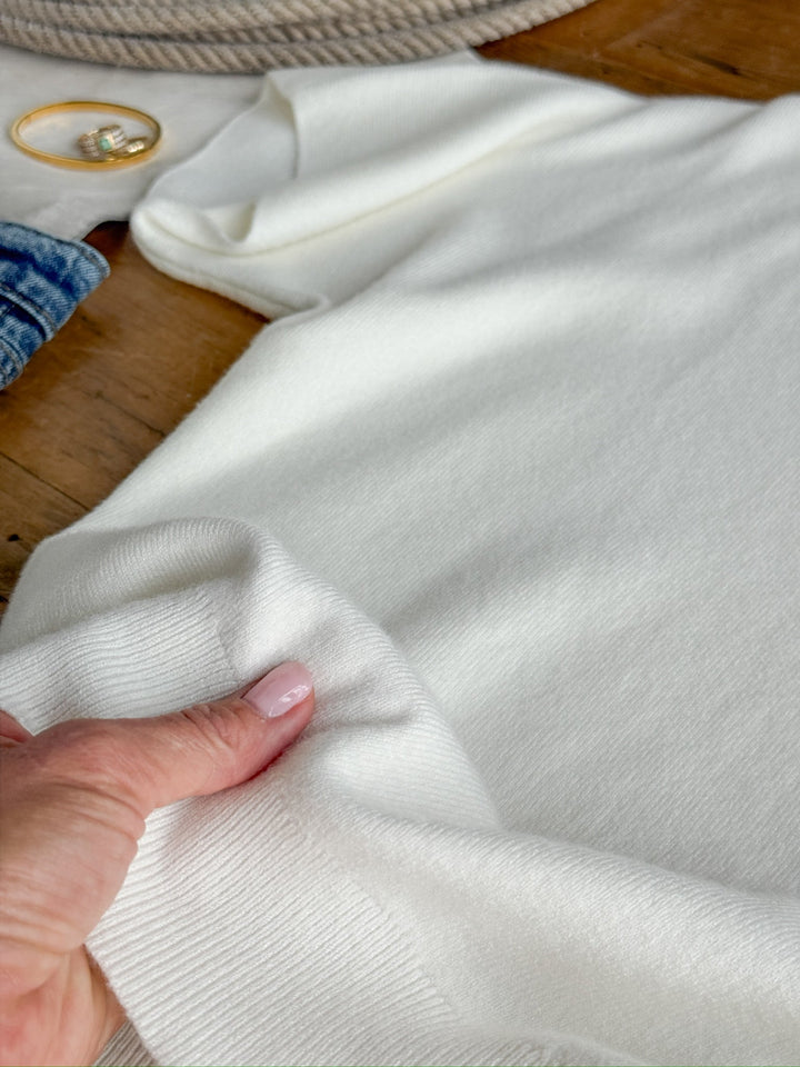 A hand with light pink nails holds the sleeve of the Panaca White Knit Top by Savannah Sevens western life{&}style on a wooden surface, with gold hoop earrings and blue denim in the background.