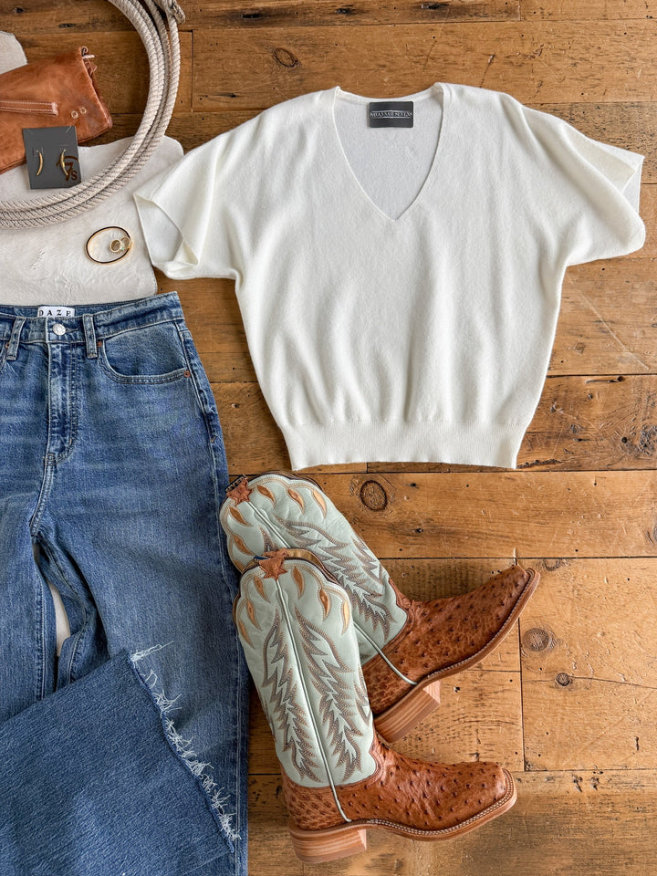 Flat lay of the Panaca White Knit Top by Savannah Sevens western life{&}style—a V-neck, short-sleeve, relaxed-fit sweater—styled with blue jeans, light blue and tan cowboy boots, a brown belt, a watch, and a brown leather bag on a wooden floor.