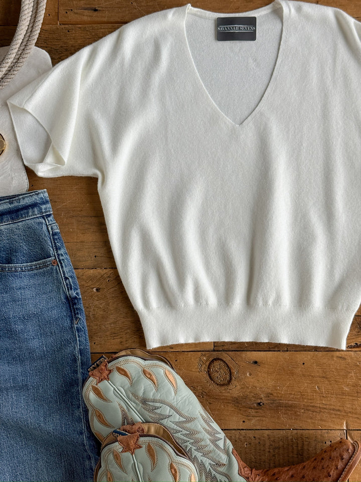 The Panaca White Knit Top by Savannah Sevens western life{&}style, paired with blue jeans, a rope-handled cream tote, and embroidered shoes, are arranged on a wooden surface.