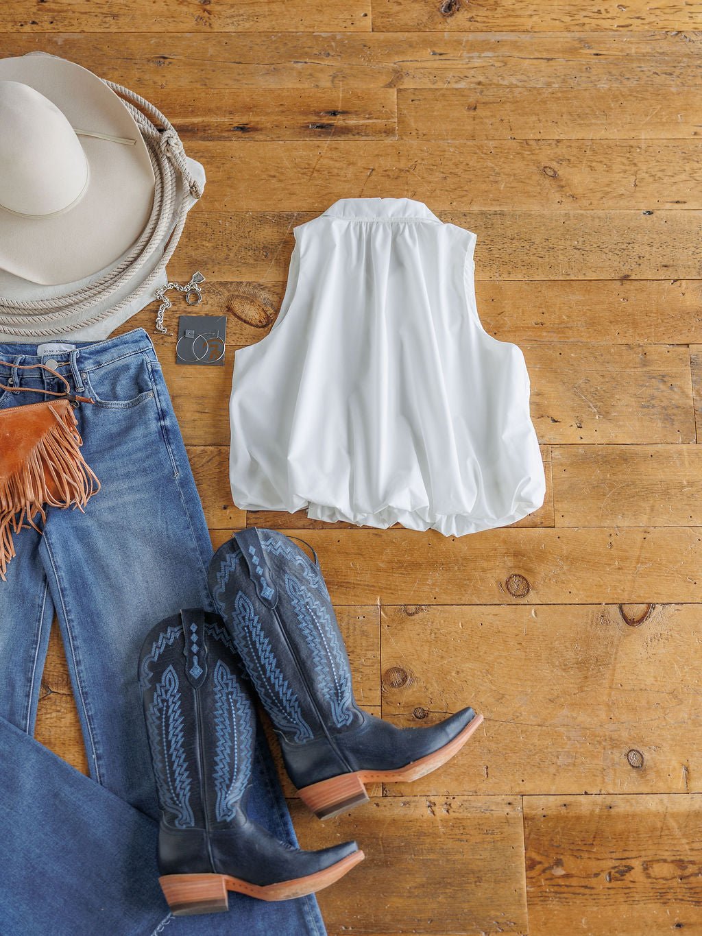 Flat lay of The Panora by Savannah Sevens western life{&}style: a sleeveless white top styled with blue fringe jeans, black and blue cowboy boots, a white cowboy hat, and silver hoop earrings on a wooden floor.
