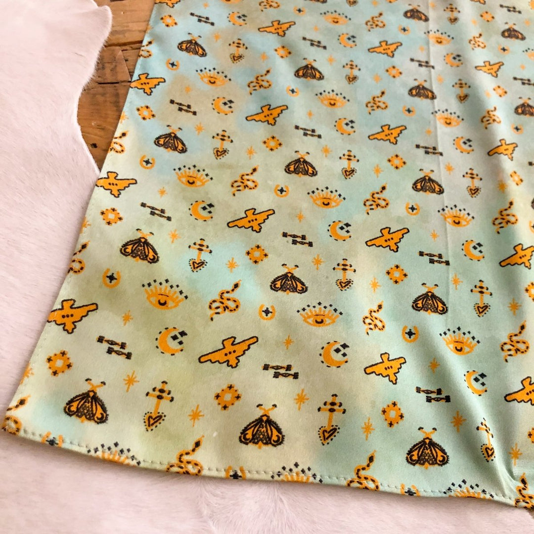 The Paoli Wild Rag by Savannah Sevens western life{&}style features a light blue-green background with yellow moths, crescent moons, stars, suns, and geometric shapes—perfect as a silk scarf or Southwest wild rag.