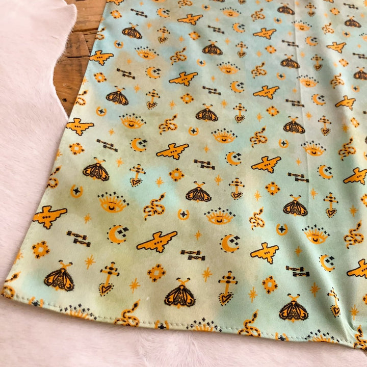 The Paoli Wild Rag by Savannah Sevens western life{&}style features a light blue-green background with yellow moths, crescent moons, stars, suns, and geometric shapes—perfect as a silk scarf or Southwest wild rag.