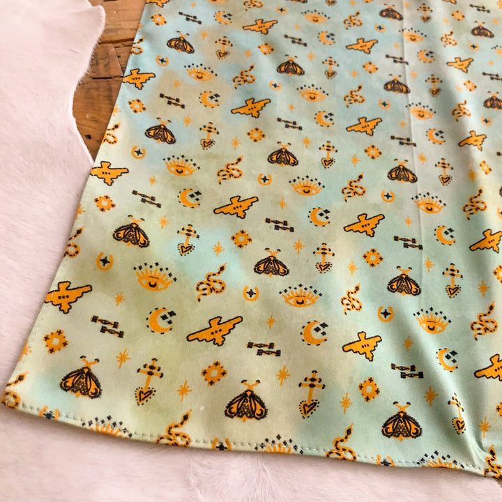 The Paoli Wild Rag by Savannah Sevens western life{&}style features a light blue-green background with yellow moths, crescent moons, stars, suns, and geometric shapes—perfect as a silk scarf or Southwest wild rag.
