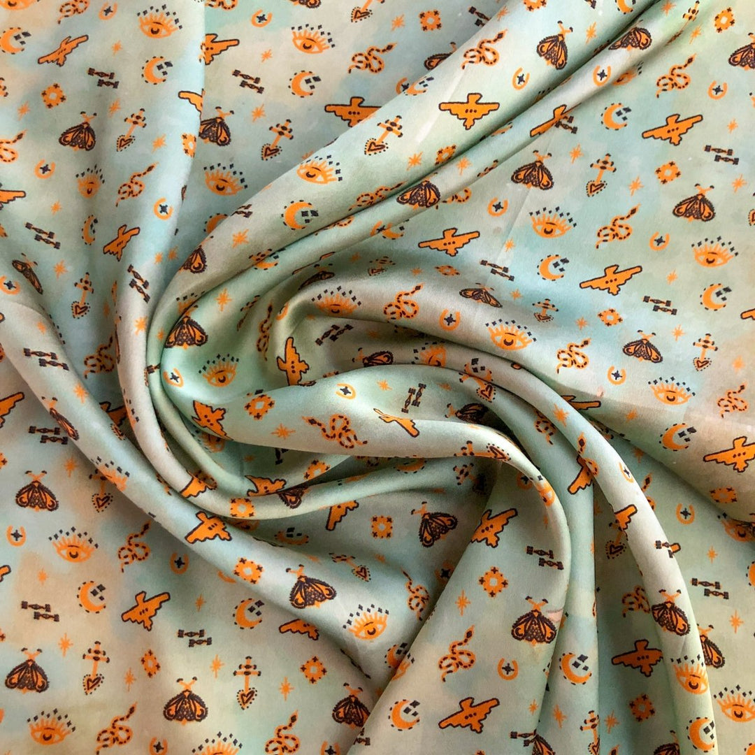 The Paoli Wild Rag by Savannah Sevens western life{&}style is a light blue silk scarf with orange stars, moons, suns, flowers, butterflies, and small dots. Its gently gathered creases create a playful, celestial-themed Southwest design.