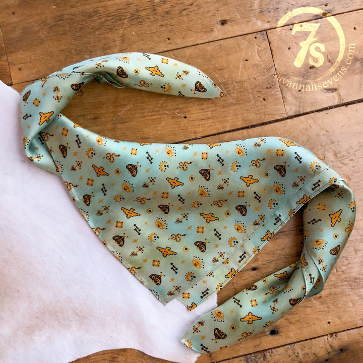 The Paoli Wild Rag by Savannah Sevens western life{&}style, a light green silk scarf with yellow and brown patterns, rests on a wooden surface over white fabric. A circular savannahsevens.com logo in the top right highlights its Southwest wild rag style.