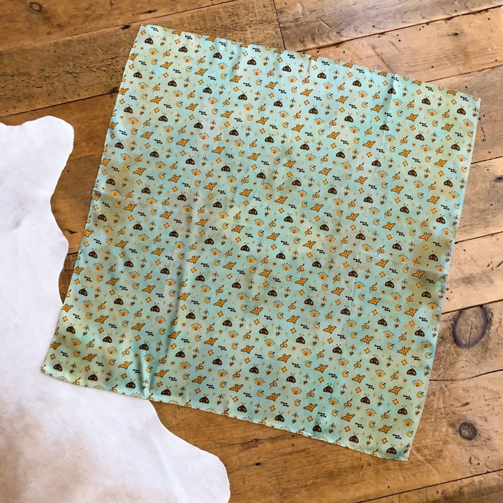 A piece of green fabric with a colorful floral print, the Paoli Wild Rag by Savannah Sevens western life{&}style, is laid flat on a wooden floor beside a white rug—perfect for styling as a Southwest wild rag or silk scarf.