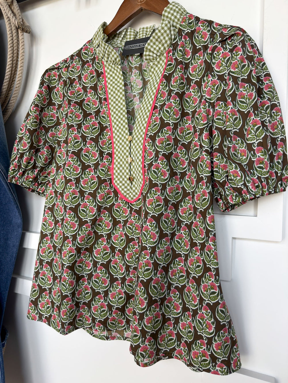 The Parada Floral Puff Sleeve Top by Savannah Sevens western life{&}style is a short-sleeved cotton blouse featuring a green, pink, and white floral pattern, a gingham ruffle neckline, and a green-checkered collar with pink trim, displayed on a wooden hanger.