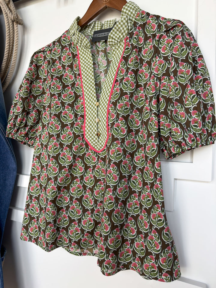 The Parada Floral Puff Sleeve Top by Savannah Sevens western life{&}style is a short-sleeved cotton blouse featuring a green, pink, and white floral pattern, a gingham ruffle neckline, and a green-checkered collar with pink trim, displayed on a wooden hanger.