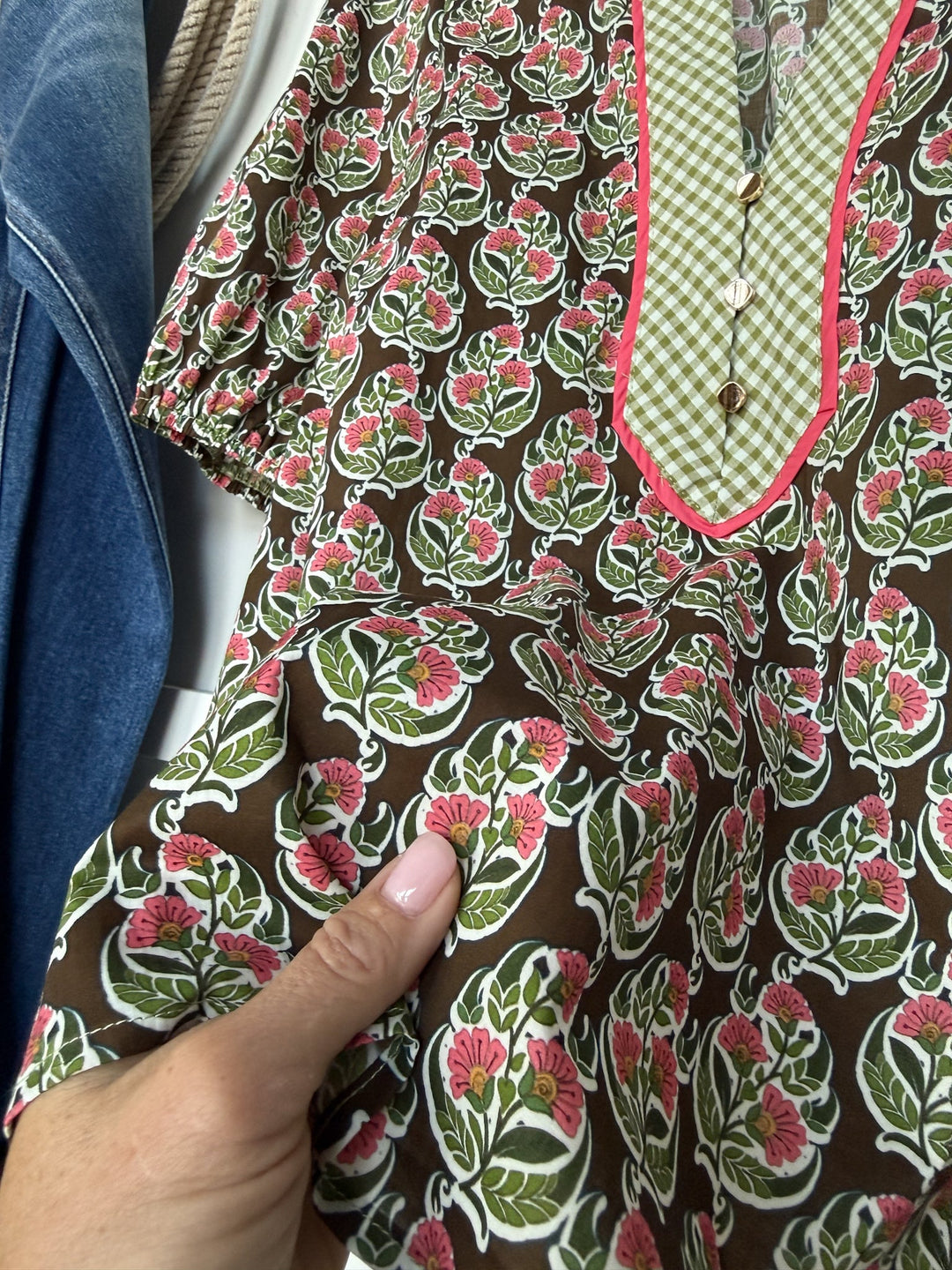 A hand holds the Parada Floral Puff Sleeve Top by Savannah Sevens western life{&}style, a cotton blouse featuring a brown, green, and pink floral pattern, a green checkered panel with buttons, and pink trim. In the background, a blue denim jacket sleeve is visible.
