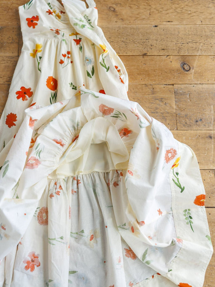 Two Park Floral Dresses by Savannah Sevens western life{&}style, featuring vibrant red, orange, and yellow flowers with green leaves on white cotton, are displayed on a wooden floor. These sleeveless midi dresses are made from light, airy fabric.