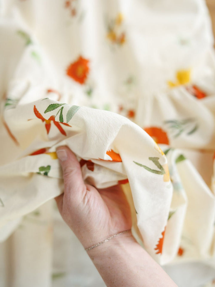 A hand holds a piece of the Park Floral Dress fabric from Savannah Sevens western life{&}style, featuring an off-white background with orange, green, and yellow spring floral prints; more of the cotton sleeveless dress shows in the background.