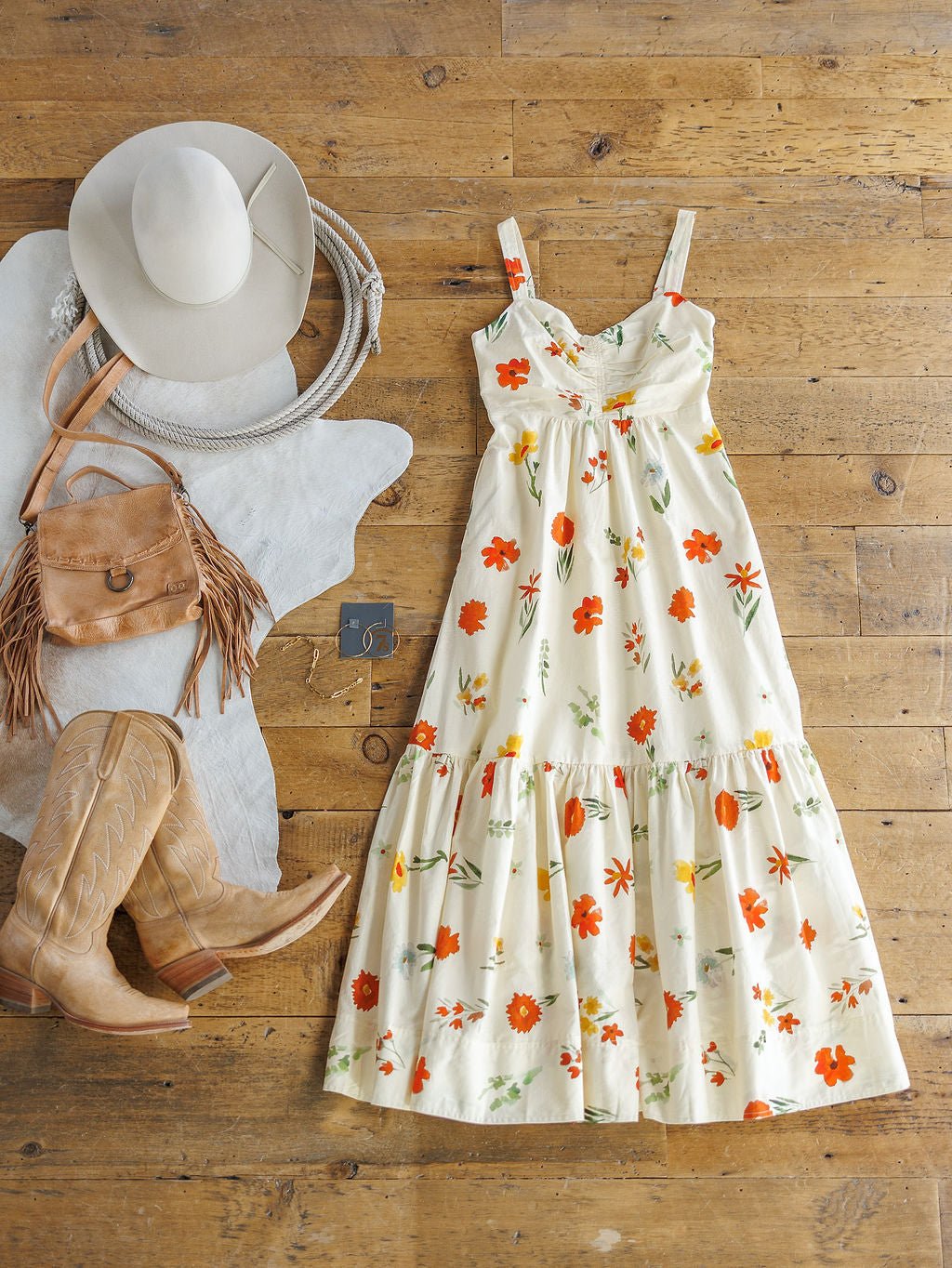 The Park Floral Dress by Savannah Sevens western life{&}style is displayed on a wooden floor with tan cowboy boots, a fringed brown leather bag, a white cowgirl hat, a coiled rope, and a white cowhide rug.
