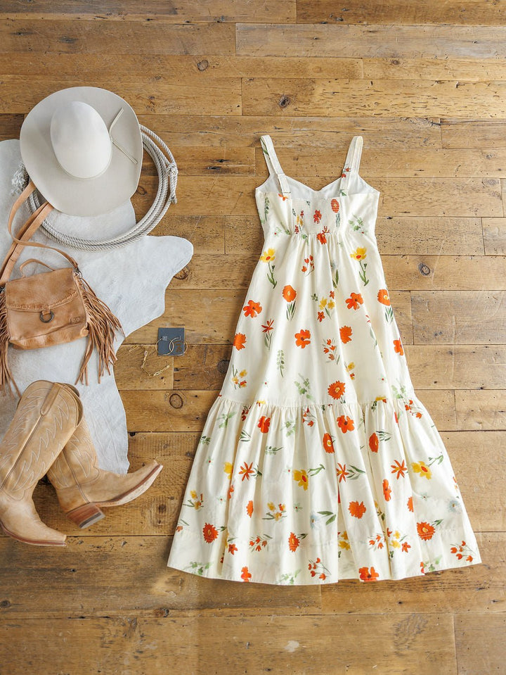 The Park Floral Dress by Savannah Sevens western life{&}style—a white cotton sleeveless dress with orange and yellow floral print—is styled with a white cowboy hat, tan fringed bag, tan boots, and a light denim jacket on a wooden floor.