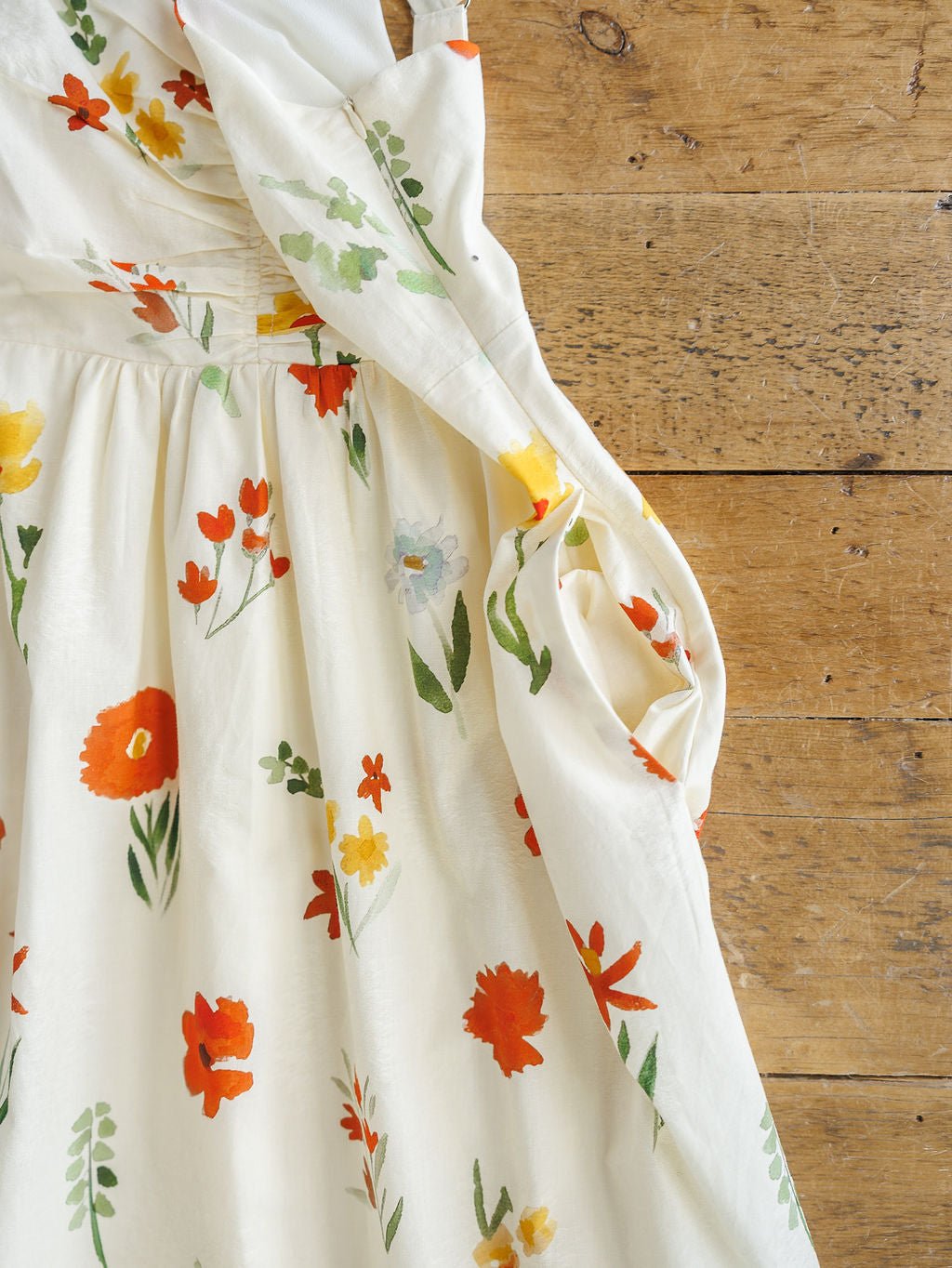 The Park Floral Dress by Savannah Sevens western life{&}style features a vibrant spring floral print on white cotton. This sleeveless dress, shown with gentle folds on rustic wood, has a light, summery look with red, orange, yellow, and green flowers.