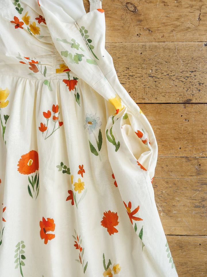 The Park Floral Dress by Savannah Sevens western life{&}style features a vibrant spring floral print on white cotton. This sleeveless dress, shown with gentle folds on rustic wood, has a light, summery look with red, orange, yellow, and green flowers.