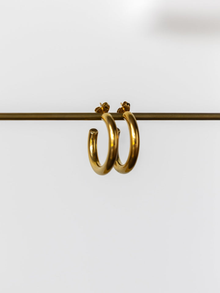 The Parton Gold Hoop Earrings by Savannah Sevens western life{&}style feature shiny, tarnish-resistant hoops with butterfly backings, displayed hanging on a thin rod against a plain white background.