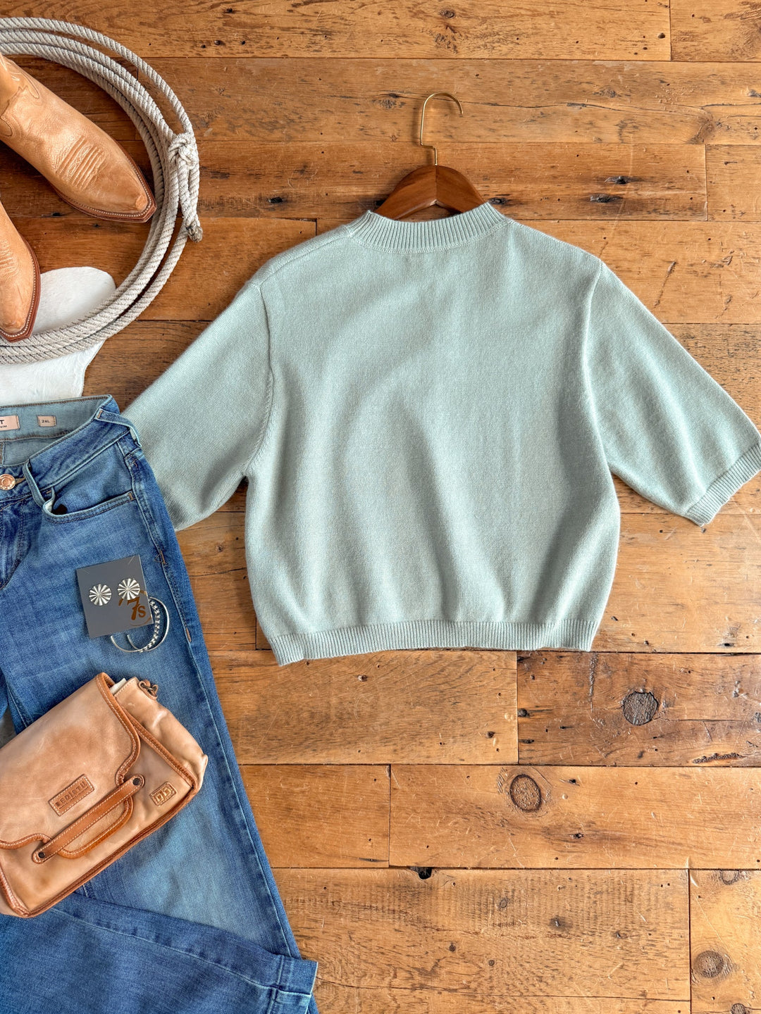 The Pasadena Sage Knit Top by Savannah Sevens western life{&}style, a relaxed fit short-sleeve sweater in light blue, is displayed on a wooden hanger and set on a wooden floor alongside blue jeans, tan shoes, and a coordinating tan purse.