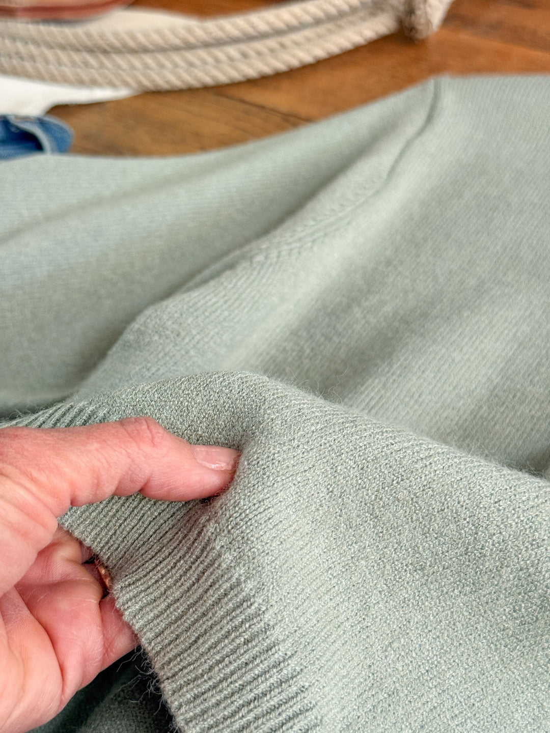 A hand gently pinches the cuff of the Pasadena Sage Knit Top by Savannah Sevens western life{&}style, highlighting its soft, textured knit fabric in a light sage green hue. This luxury sweater offers a relaxed fit, with rope and wooden surfaces visible in the background.