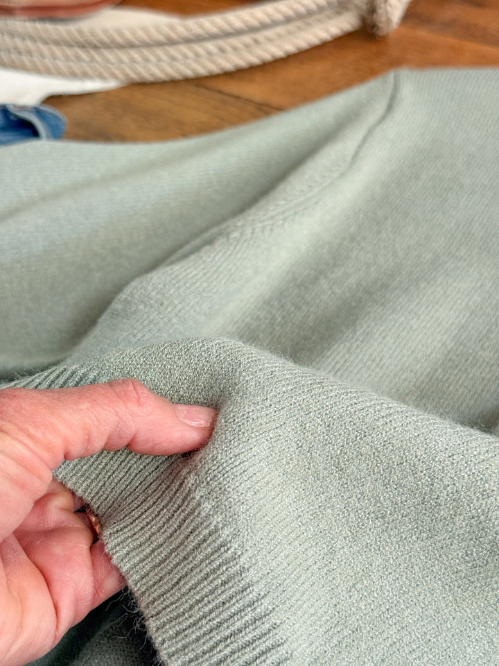 A hand gently pinches the cuff of the Pasadena Sage Knit Top by Savannah Sevens western life{&}style, highlighting its soft, textured knit fabric in a light sage green hue. This luxury sweater offers a relaxed fit, with rope and wooden surfaces visible in the background.