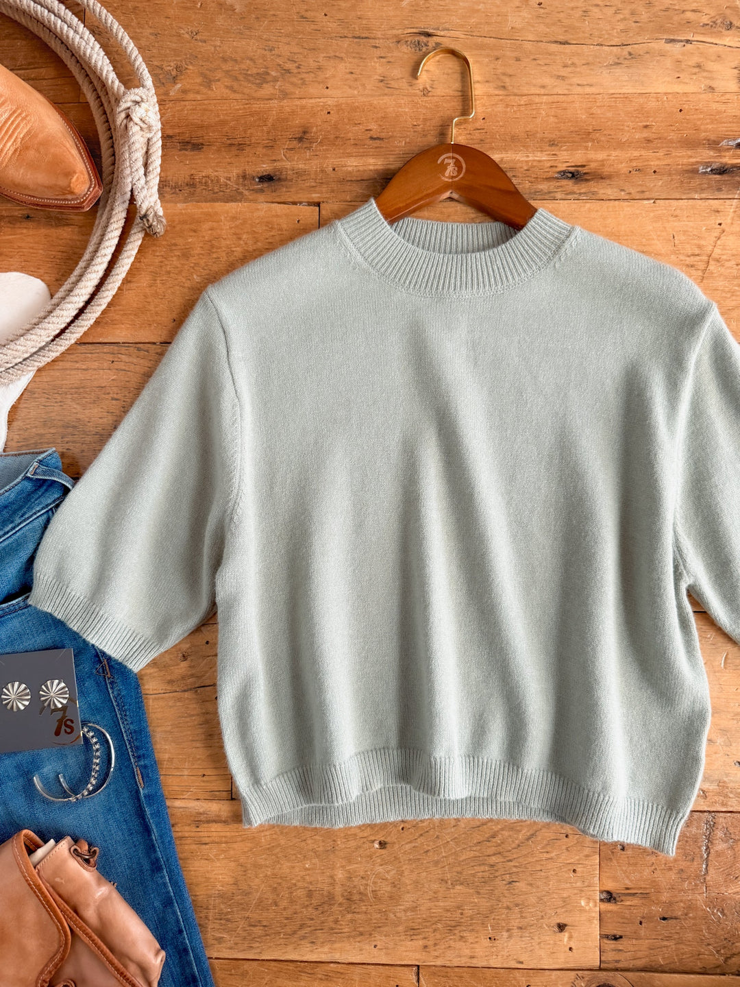 The Pasadena Sage Knit Top by Savannah Sevens western life{&}style is a luxurious, light gray knit featuring a relaxed fit and short sleeves. Presented on a hanger and laid flat on a wooden floor, it’s styled with a denim jacket, brown leather bag, and rope.