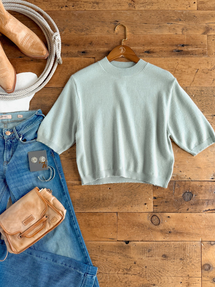 A Pasadena Sage Knit Top by Savannah Sevens western life{&}style in a light blue hue with short sleeves is displayed on a hanger, laid on a wooden floor and surrounded by jeans, a tan purse, tan boots, and daisy-themed jewelry.