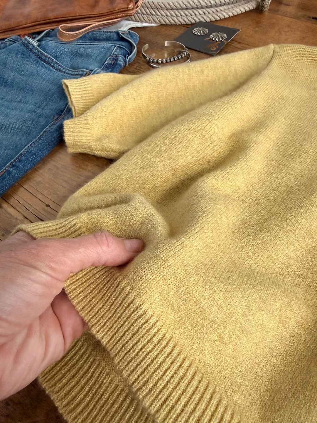 A hand holds the hem of the Pasadena Yellow Chartreuse Knit Top by Savannah Sevens western life{&}style, displayed on a wooden surface alongside blue jeans, a brown bag, a silver bracelet, and earrings.