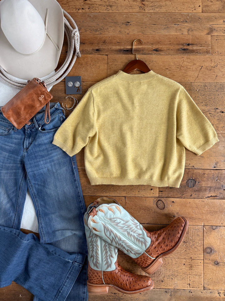 Flat lay of the Savannah Sevens western life{&}style Pasadena Yellow Chartreuse Knit Top, blue jeans, tan cowboy boots with light blue stitching, a white cowboy hat, a rope, brown leather bag, and silver earrings on a wooden floor.