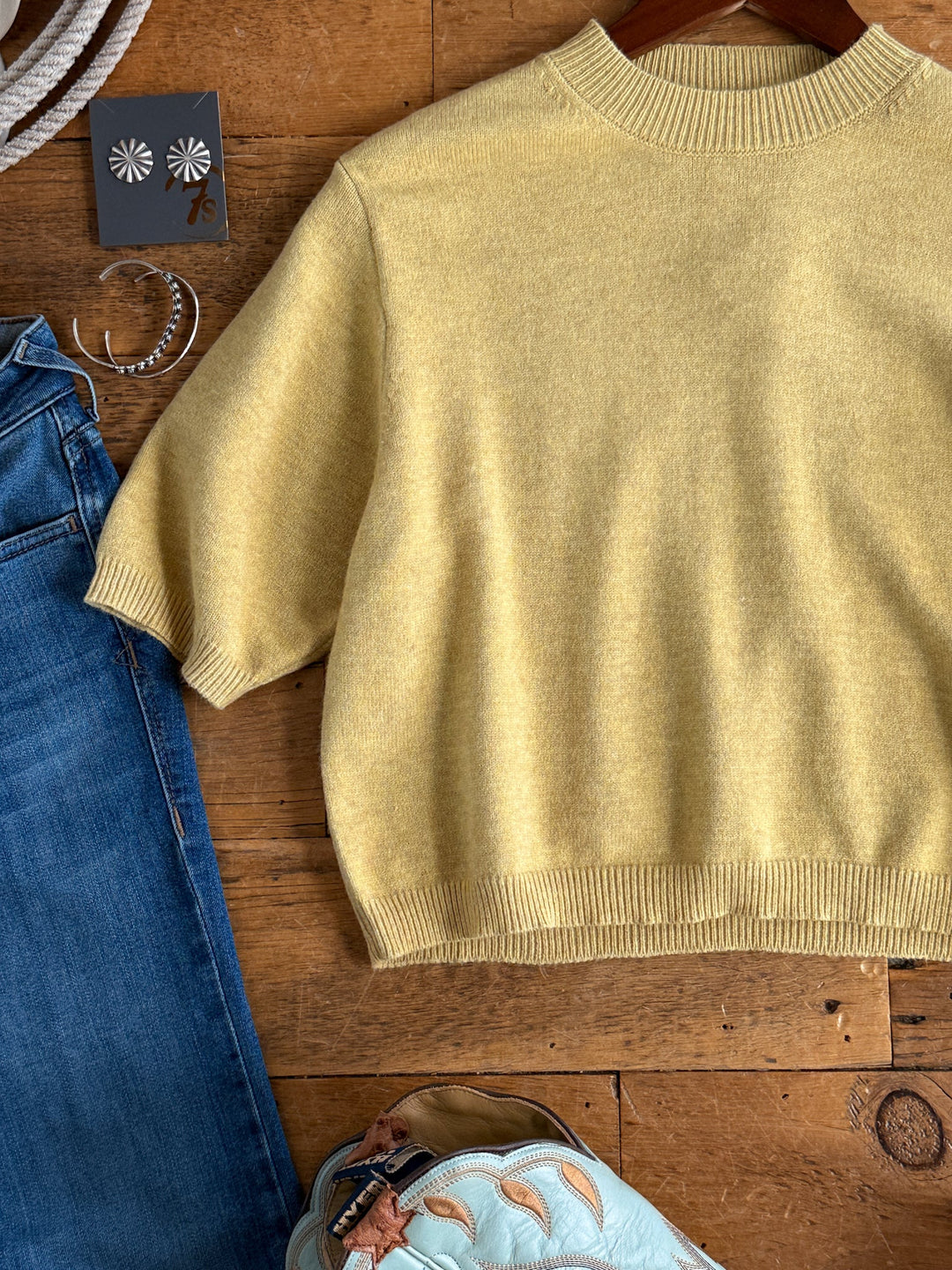 The Pasadena Yellow Chartreuse Knit Top by Savannah Sevens western life{&}style is styled with blue jeans, floral earrings, a silver bracelet, a light blue purse, and rope detail, all arranged on a wooden surface.