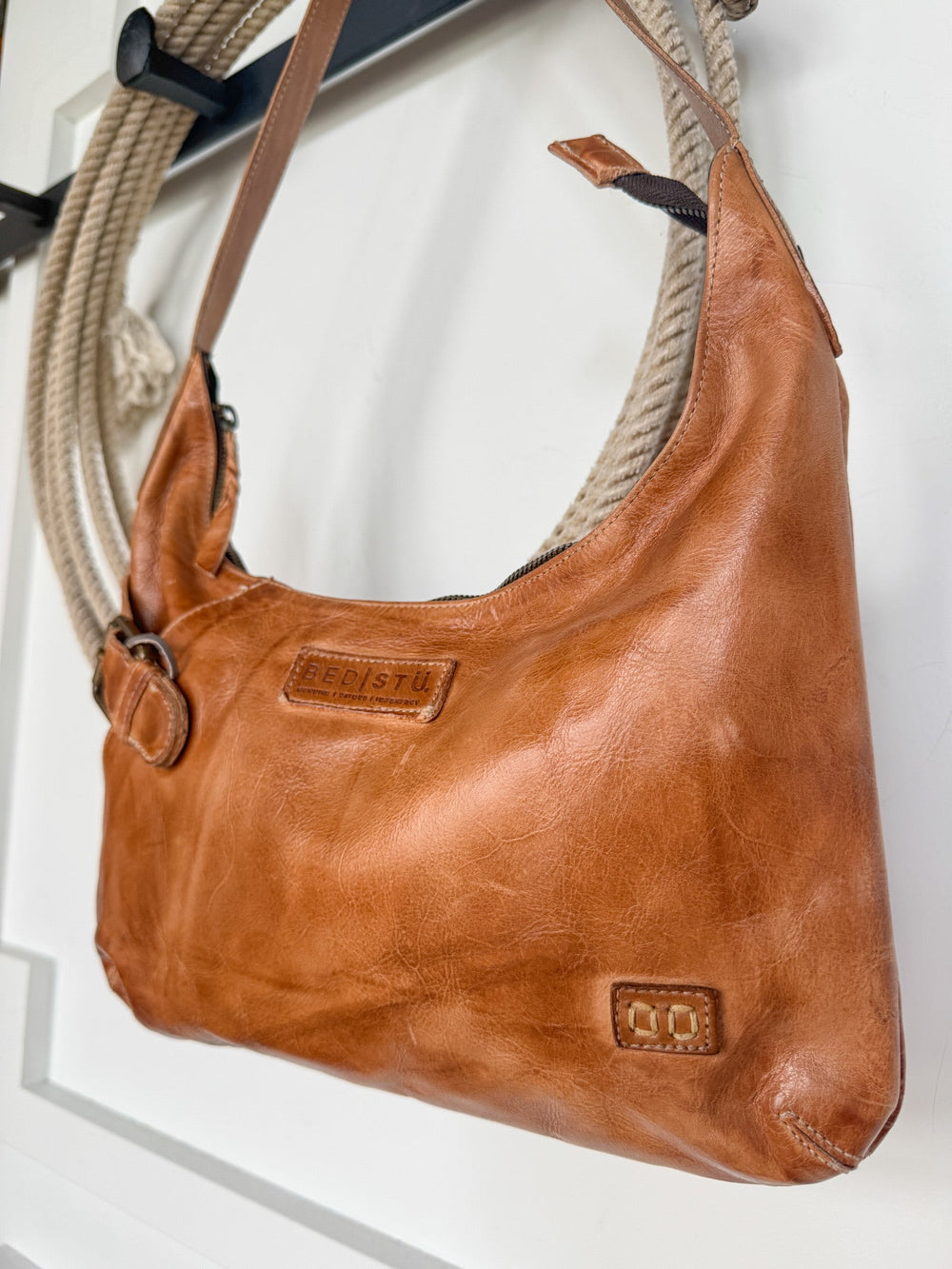 A pecan Pasco Leather Handbag from Savannah Sevens western life{&}style, featuring a zipper closure, visible stitching, and brand embossing, hangs on a hook with a coiled rope in the background.