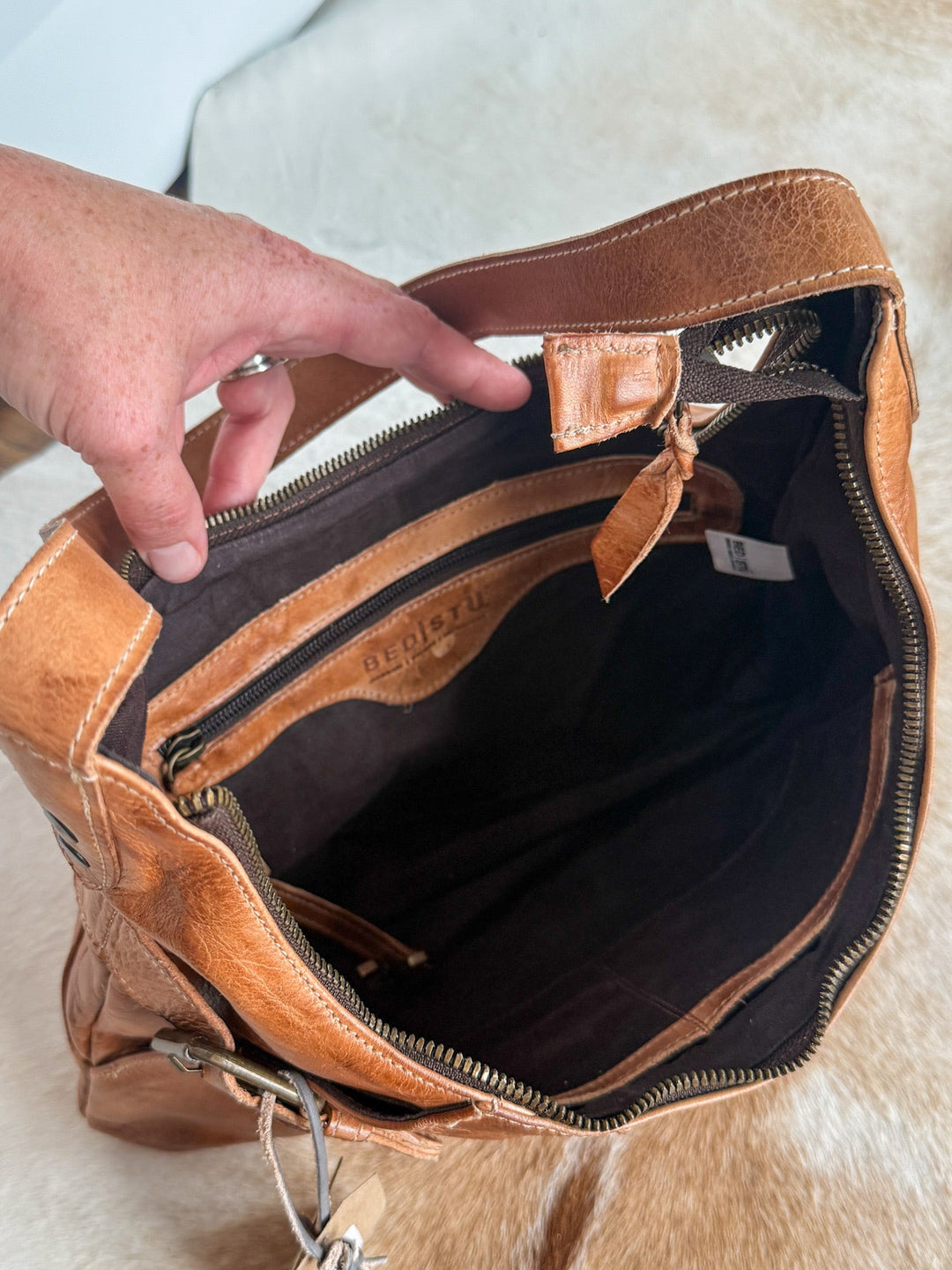 A hand holds open a Savannah Sevens western life{&}style Pasco Leather Handbag, revealing its spacious, dark-lined interior and an inner zippered pocket, set on a soft, light-colored surface.