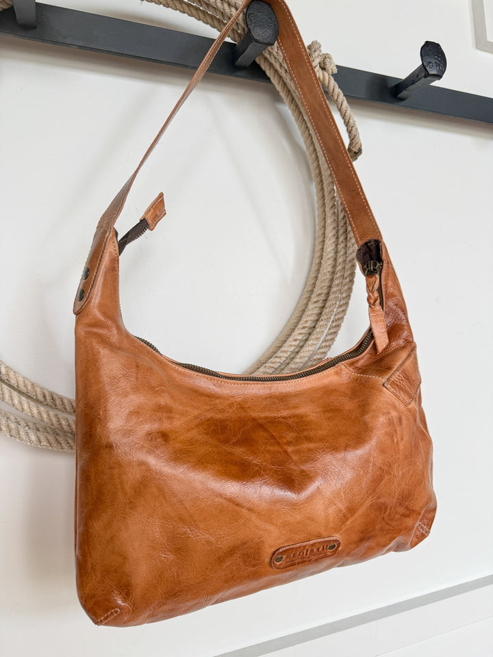 A Pasco Leather Handbag by Savannah Sevens western life{&}style, featuring a pecan leather finish and zipper closure, hangs on a black wall hook. In the background, a coiled rope rests against a white wall.