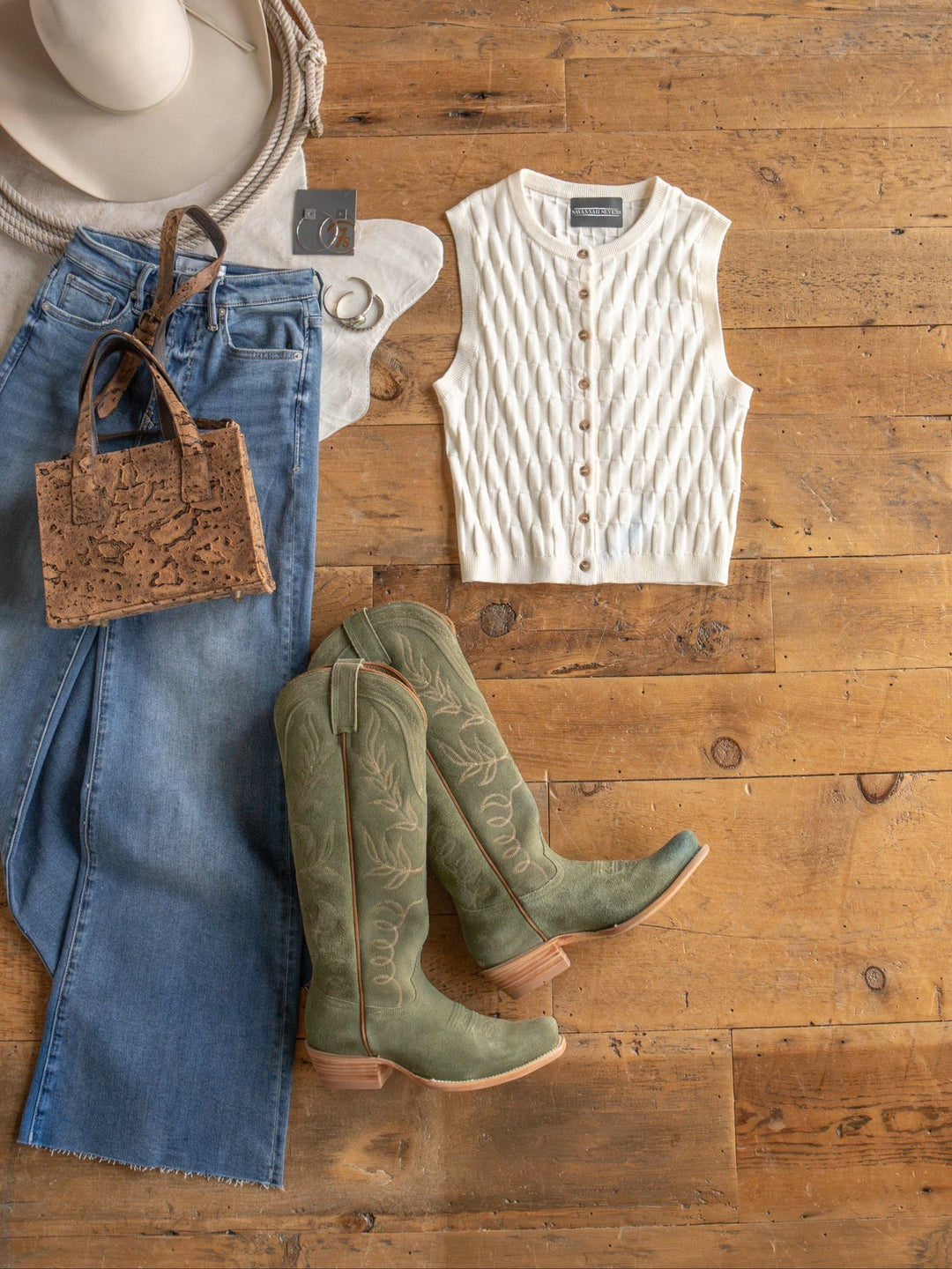 Flat lay of The Patero, a textured sleeveless knit top by Savannah Sevens western life{&}style, paired with jeans, sage green cowboy boots, handbag, beige wide-brim hat, and silver hoop earrings on a wooden floor.