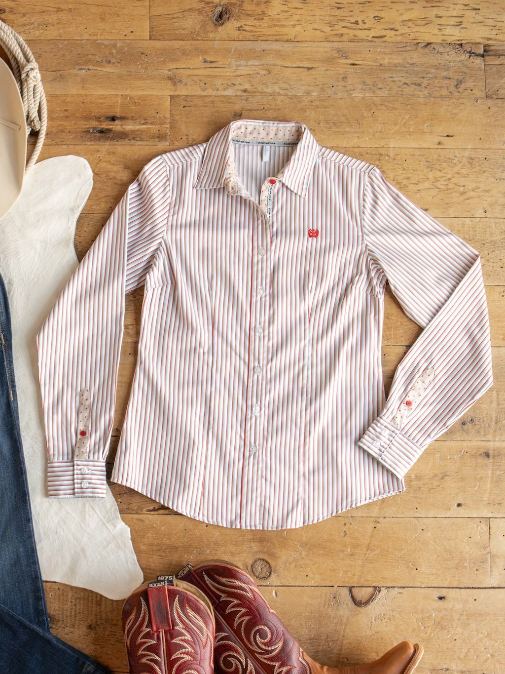 The Patton Striped Tencel Performance Shirt by Savannah Sevens western life{&}style, featuring red and white vertical stripes, is laid flat on a wooden floor alongside a cowboy boot, jeans, and a piece of white fabric. This women's long sleeve top combines classic style and comfort with its Tencel blend construction.