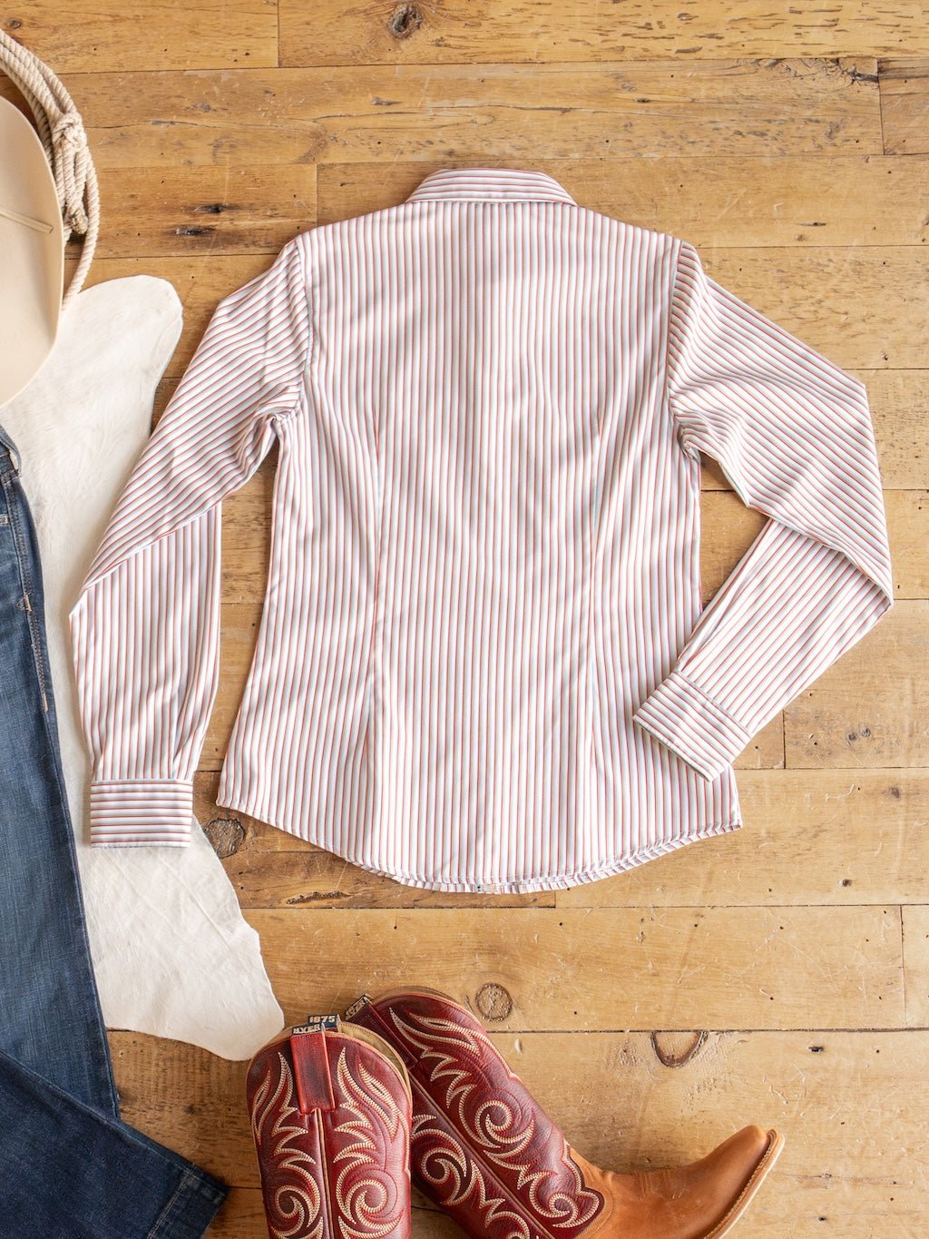 The Patton Striped Tencel Performance Shirt by Savannah Sevens western life{&}style is laid flat on a wooden floor next to a pair of red cowboy boots, a white cloth, jeans, and a beige hat.