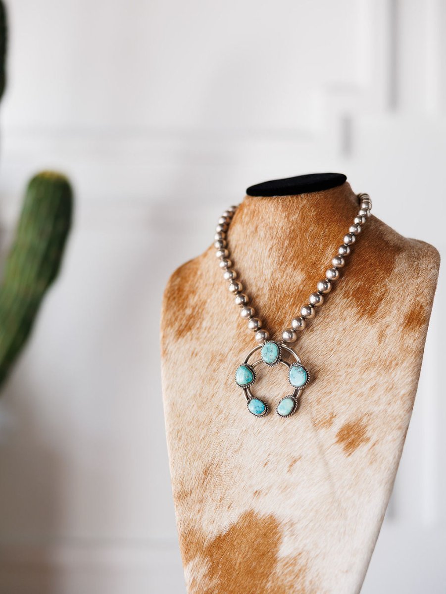 The Pawnee City Necklace by Savannah Sevens western life{&}style, featuring sterling silver beads and turquoise stones, is displayed on a cowhide-covered bust with a blurred cactus in the background, highlighting Native American handmade craftsmanship.