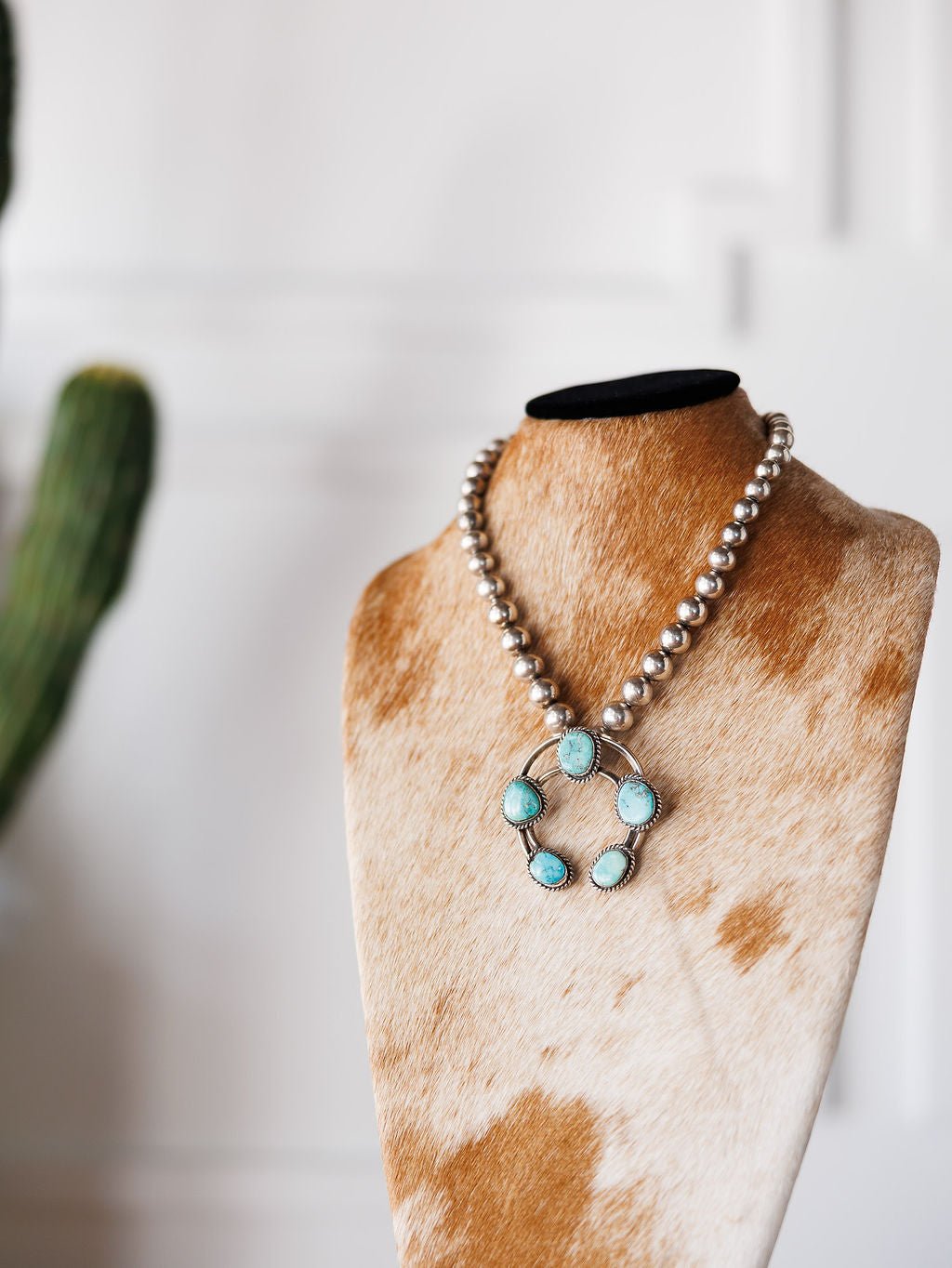 The Pawnee City Necklace by Savannah Sevens western life{&}style, featuring sterling silver beads and turquoise stones, is displayed on a cowhide-covered bust with a blurred cactus in the background, highlighting Native American handmade craftsmanship.